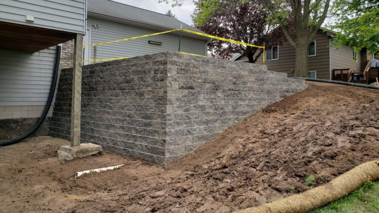 A construction worker builds a retaining wall in front of a house, with tools and materials visible on the ground.?
